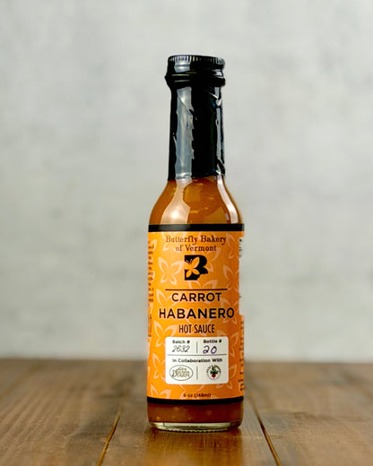 A bottle of Butterfly Bakery of Vermont Carrot Habanero Hot Sauce, featuring a bright orange label and black cap, set on a wooden table against a neutral gray background.
