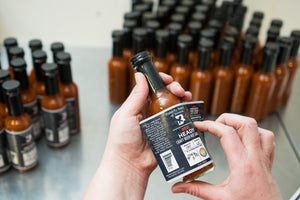 Person applying a label to a bottle of hot sauce with a blurred background of more bottles.