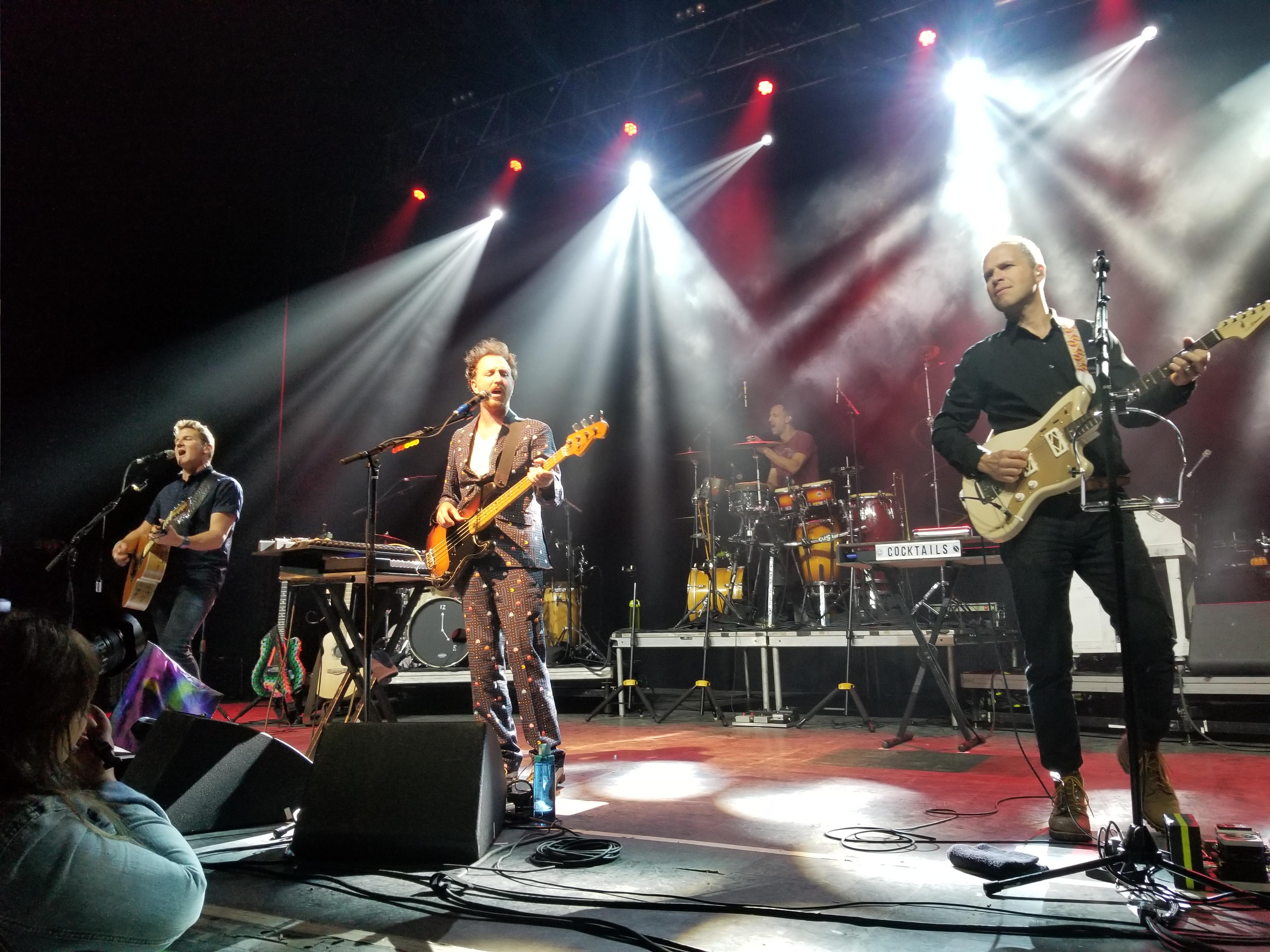 Guster performing on stage with bright lights and instruments.