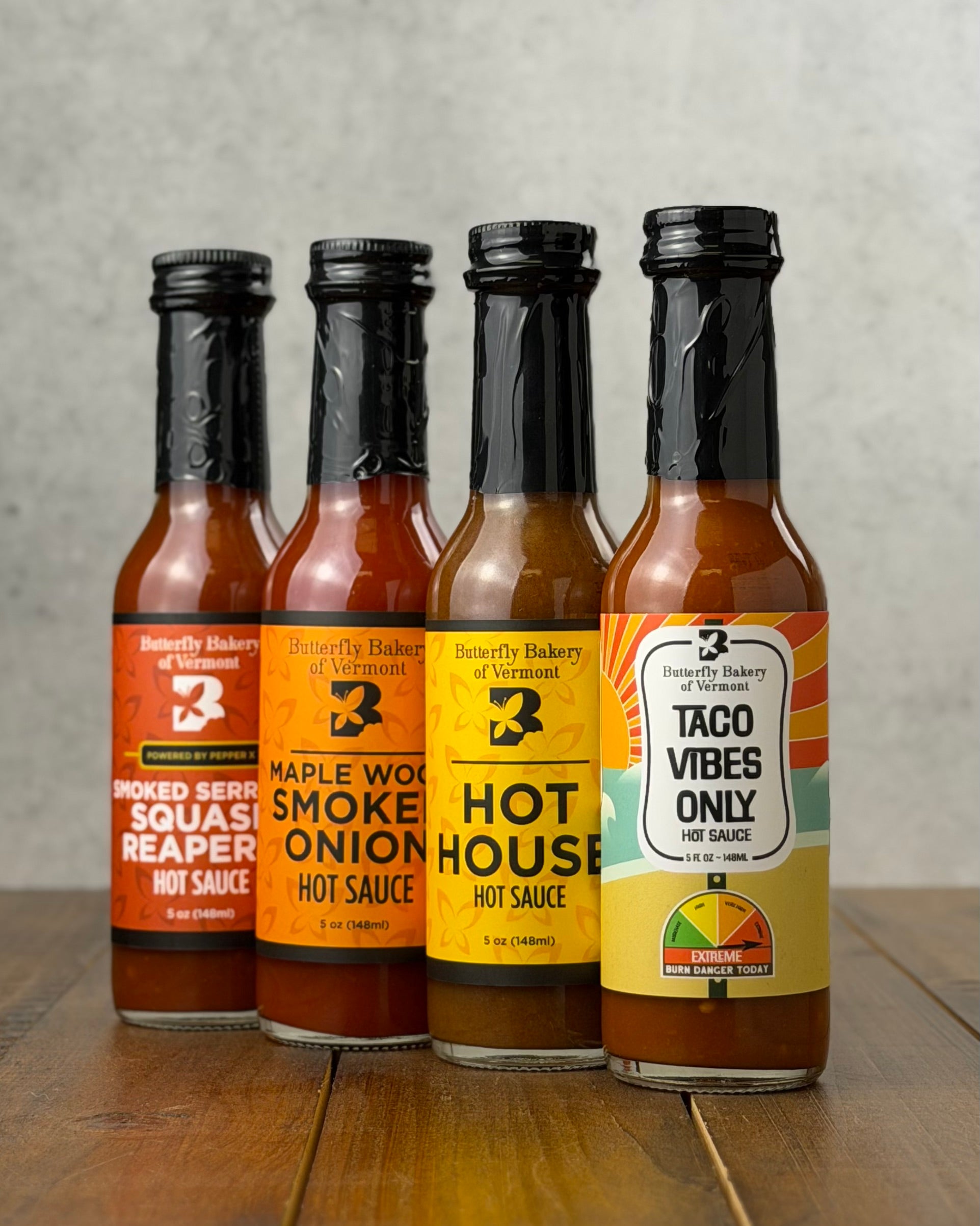 Bottles of all four hot sauces from Butterfly Bakery of Vermont that have appeared on Hot Ones, all on a wooden surface
