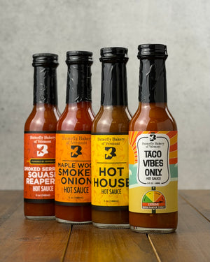 Bottles of all four hot sauces from Butterfly Bakery of Vermont that have appeared on Hot Ones, all on a wooden surface