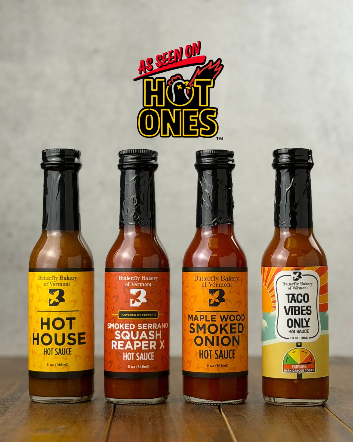 Bottles of all four hot sauces from Butterfly Bakery of Vermont that have appeared on Hot Ones, all on a wooden surface with the 'As Seen On Hot Ones' logo