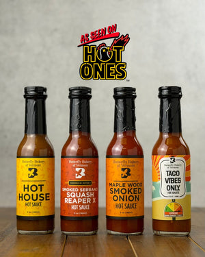 Bottles of all four hot sauces from Butterfly Bakery of Vermont that have appeared on Hot Ones, all on a wooden surface with the 'As Seen On Hot Ones' logo