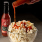 Hot sauce being poured onto popcorn with a bottle of Ooh La La hot sauce in the background.