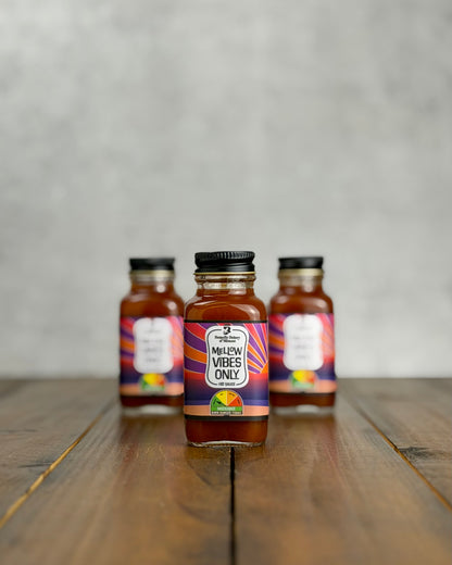 Three 2oz bottles of 'Mellow Vibes Only' sauce on a wooden surface with a gray background