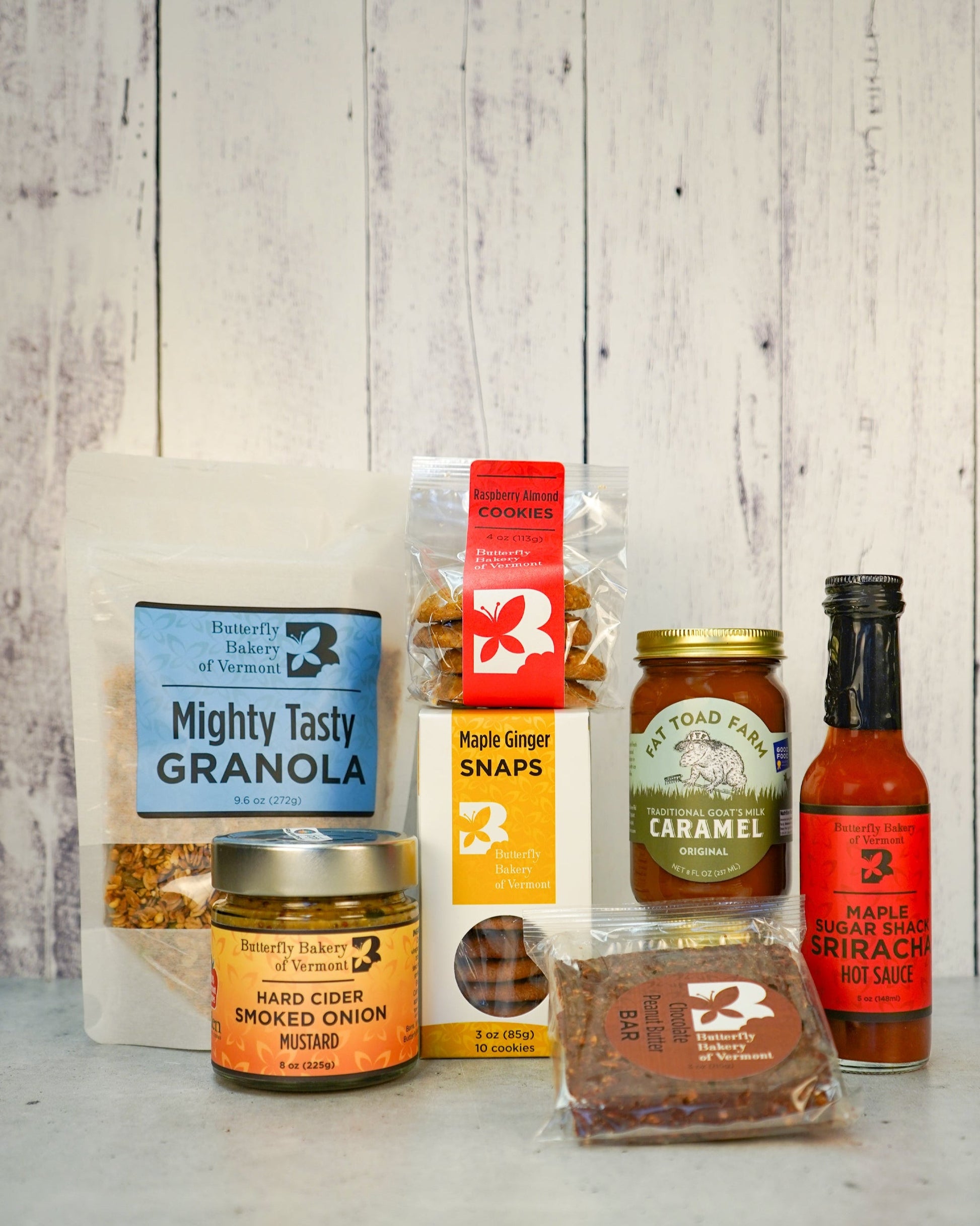 Assorted Butterfly Bakery of Vermont products including hot sauce, mustard, granola and Fat Toad Farm caramel on a stone surface with a gray background.