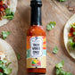 Bottle of Taco Vibes Only Hot Sauce from Butterfly Bakery of Vermont lying on a wood table alongside tacos with meat, veggies and hot sauce on them