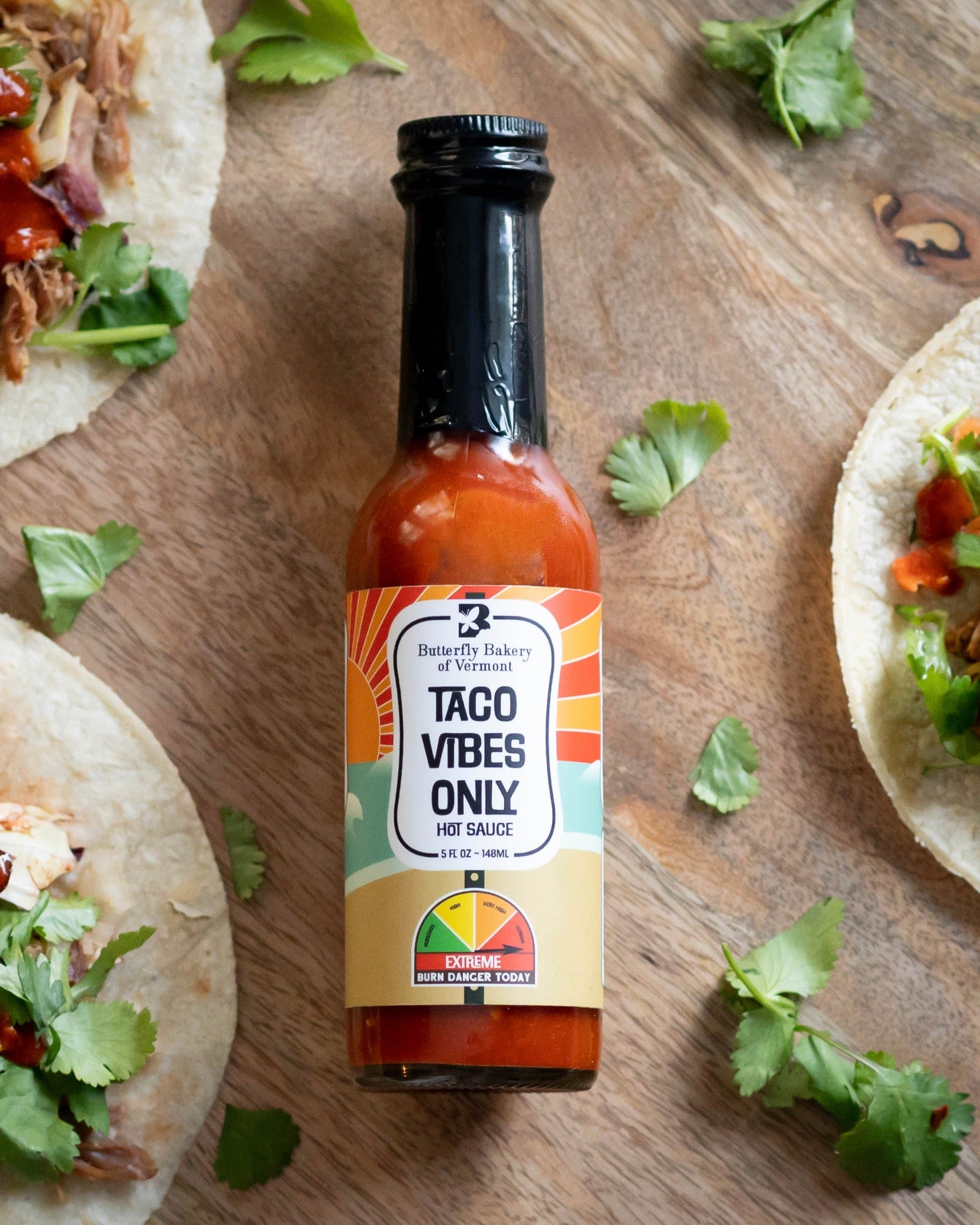 Bottle of Taco Vibes Only Hot Sauce from Butterfly Bakery of Vermont lying on a wood table alongside tacos with meat, veggies and hot sauce on them