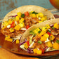 Two fish tacos with mango salsa and hot sauce on a wooden plate against a rustic background.