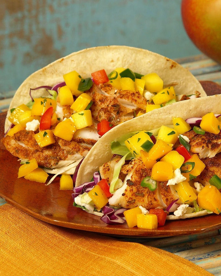 Two fish tacos with mango salsa and hot sauce on a wooden plate against a rustic background.