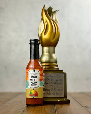 Bottle of 'Taco Vibes Only' hot sauce with a gold trophy on a wooden surface.