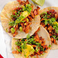 Three tacos with meat, vegetables, and hot sauce next to a lime wedge on a white plate.