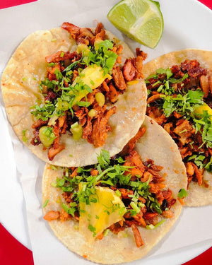Three tacos with meat, vegetables, and hot sauce next to a lime wedge on a white plate.