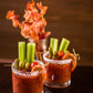 Two short glasses of bloody marys on a black table with celery, olives and pickles