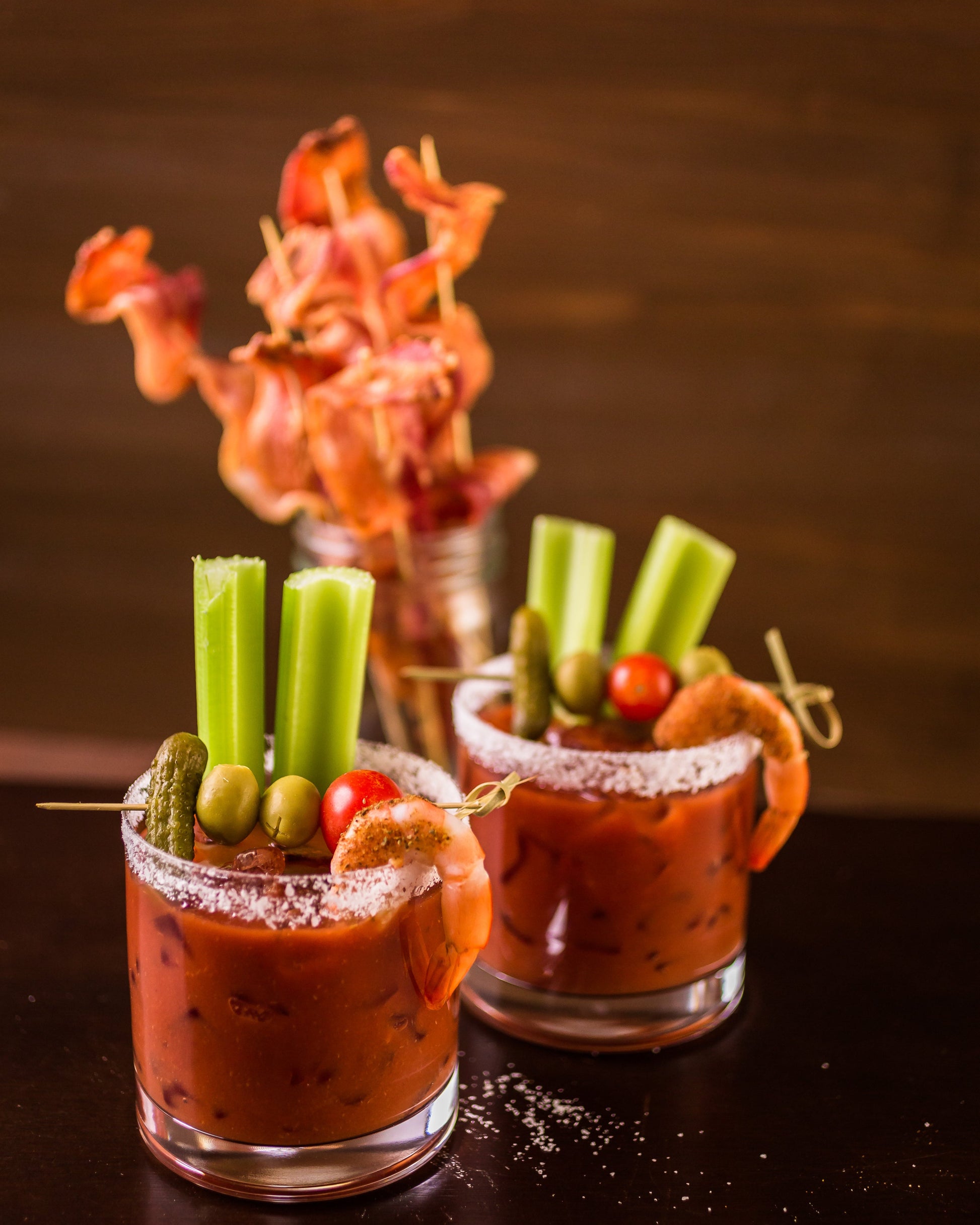 Two short glasses of bloody marys on a black table with celery, olives and pickles