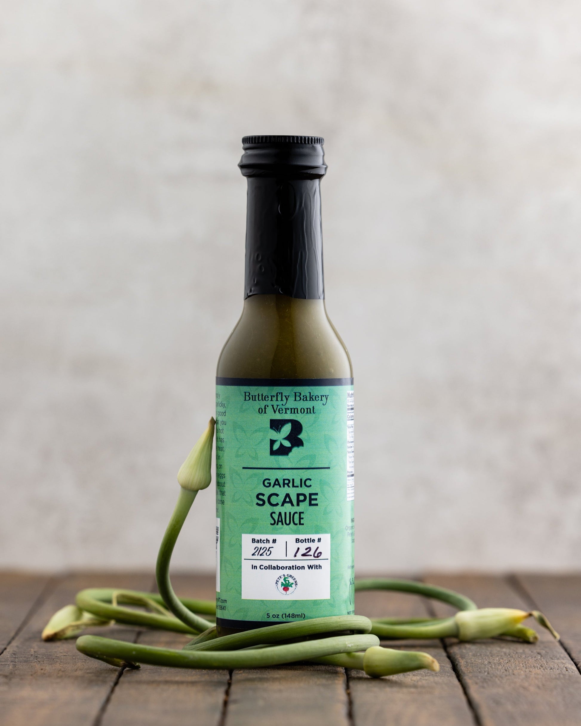 Bottle of Butterfly Bakery of Vermont Scape Sauce surrounded by garlic scapes on a wood table
