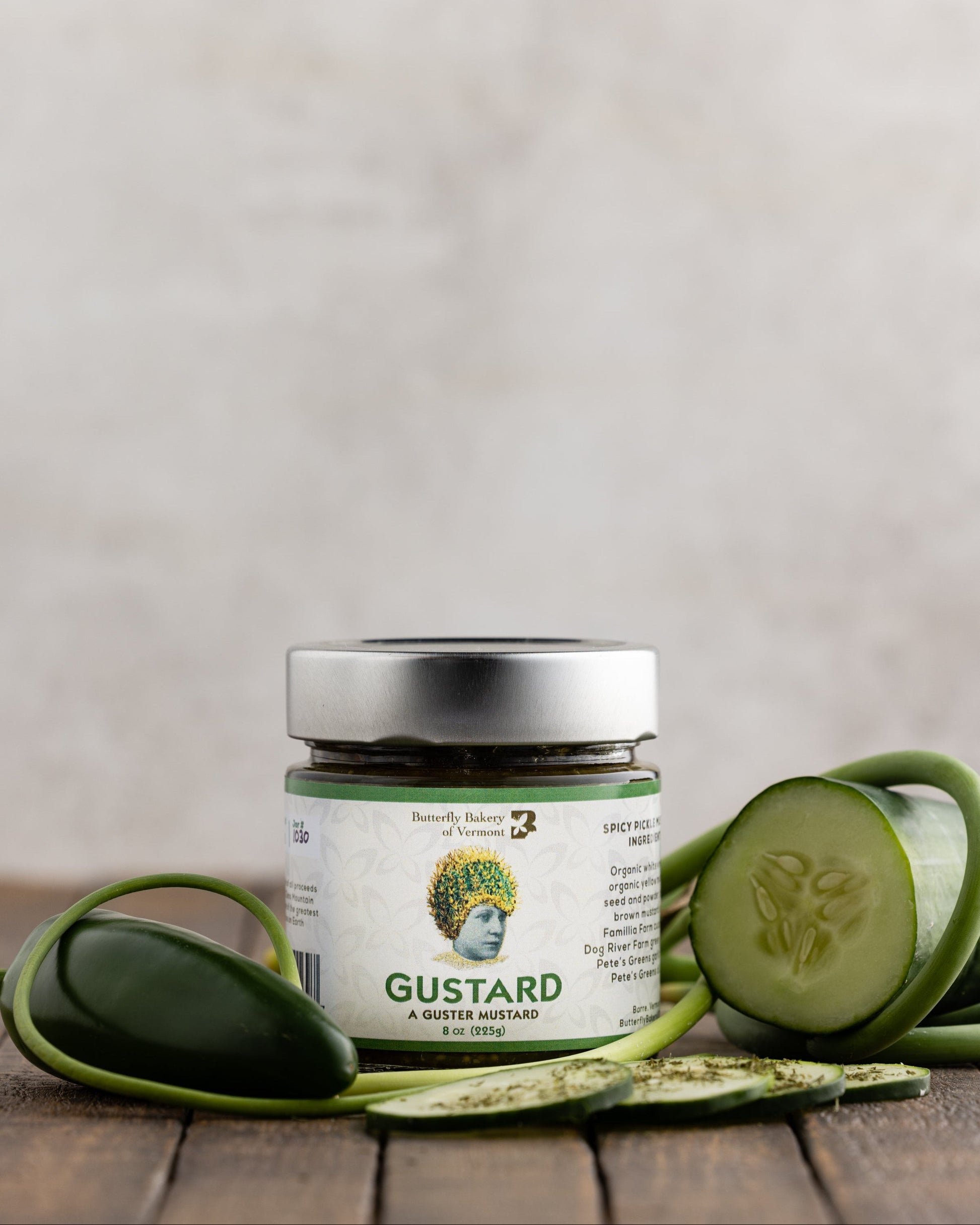 Jar of Gustard Mustard from Butterfly Bakery of Vermont on a wood surface surrounded by garlic scapes, cucumber and jalapeno